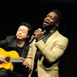 A man singing and playing an acoustic guitar next to another man.