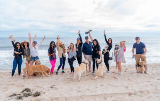 Group of people and dogs joyfully posing on a beach with ocean waves behind them.