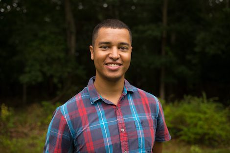 A young man in a plaid shirt standing in a wooded area.