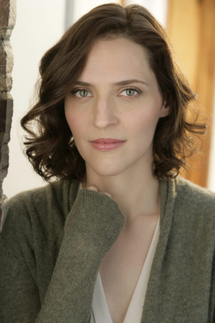 A woman in a green cardigan leaning against a brick wall.