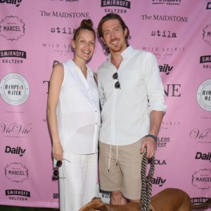 A man and woman standing next to a dog on a pink background.