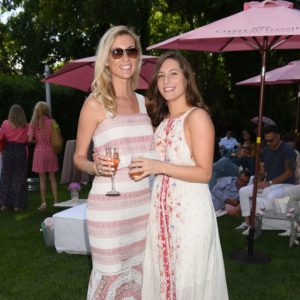 Two women standing under a pink umbrella at an outdoor event.