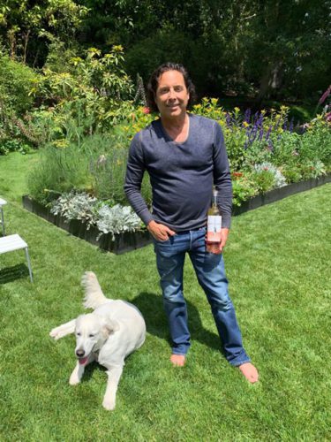 A man standing next to a dog in a garden with a bottle of wine.