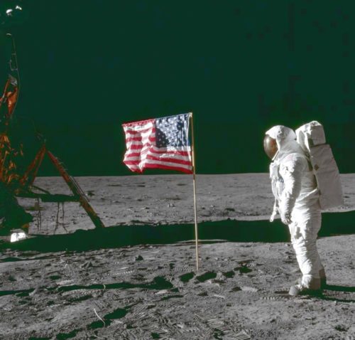 A man in a spacesuit stands next to a flag on the moon.