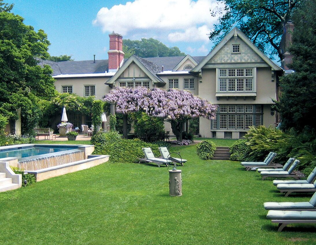 Elegant mansion with lush garden, pool, and lounging area under a blooming tree.