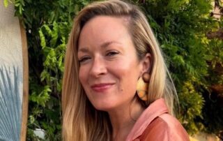 Smiling woman with long blonde hair and large wooden earrings outdoors.