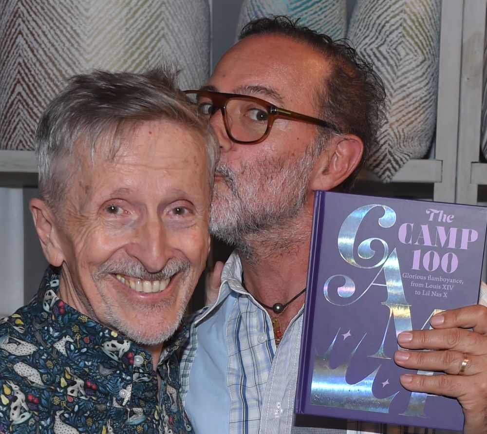 Two men smiling, one kissing the other on the cheek, holding a book.