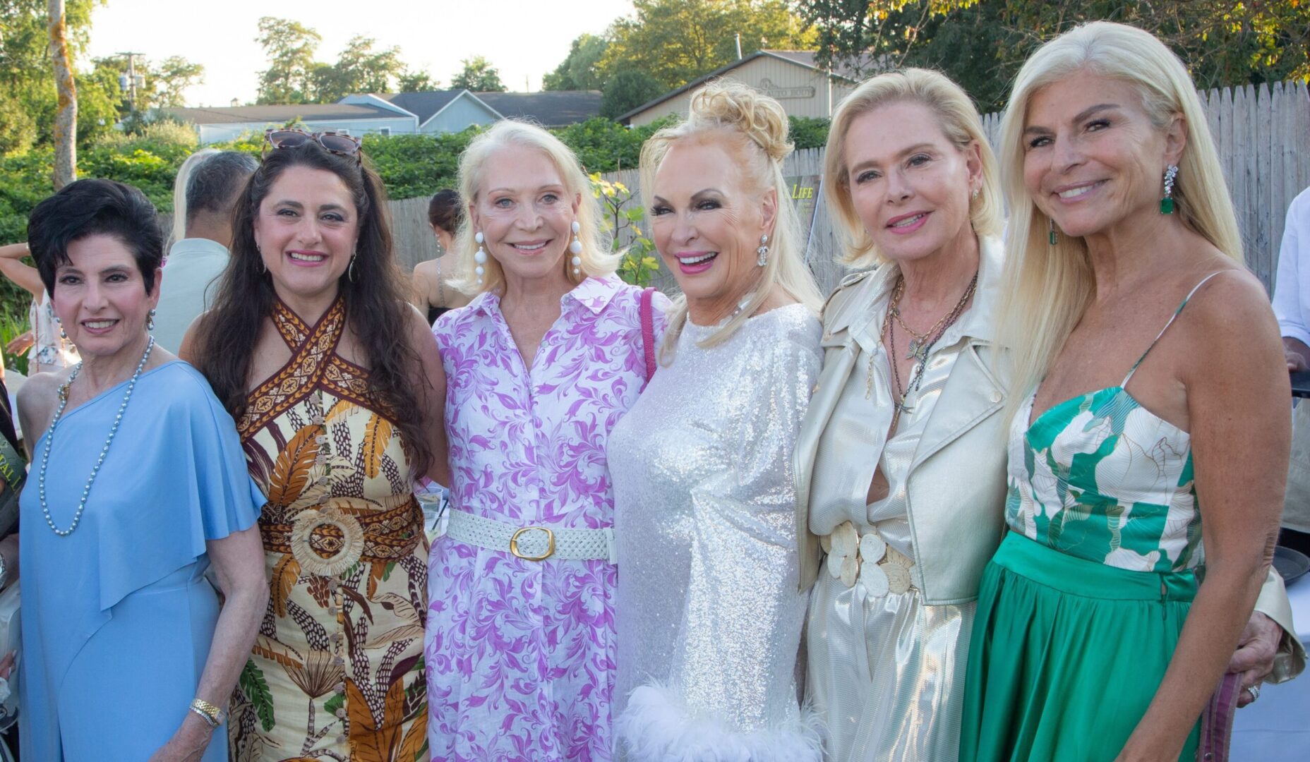 Five women smiling together at an outdoor event in stylish outfits.