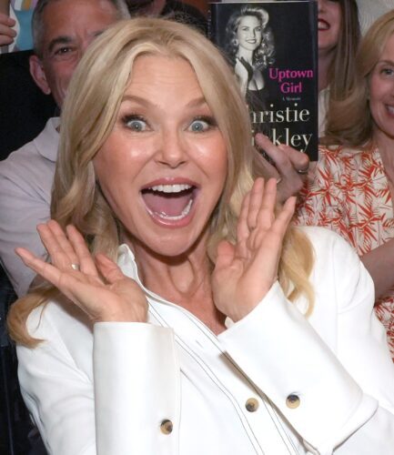 Excited woman with blonde hair smiling widely, hands near her face.