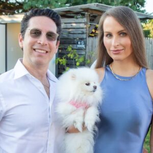 A man and woman posing outdoors with a fluffy white dog.