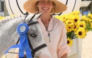 A smiling woman in a hat poses with a horse wearing a blue ribbon.
