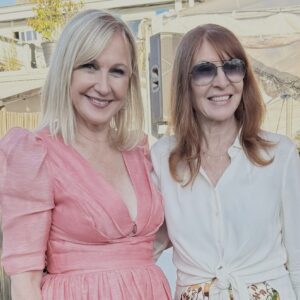 Two women smiling outdoors, one in a pink dress and the other in a white blouse with sunglasses.