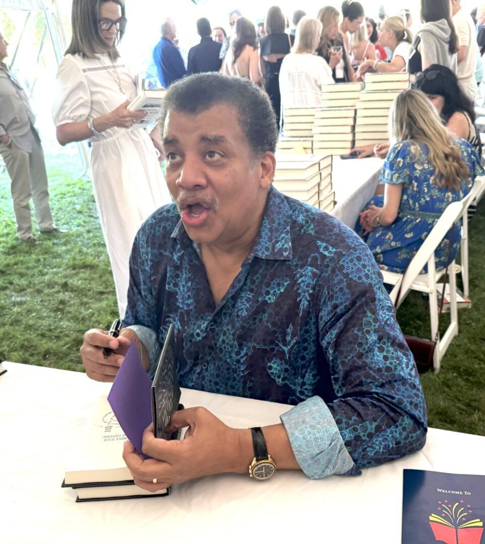Man signing a book at an outdoor event with people in the background.