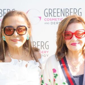 Two women wearing sunglasses posing in front of a Greenberg Cosmetic Surgery backdrop.