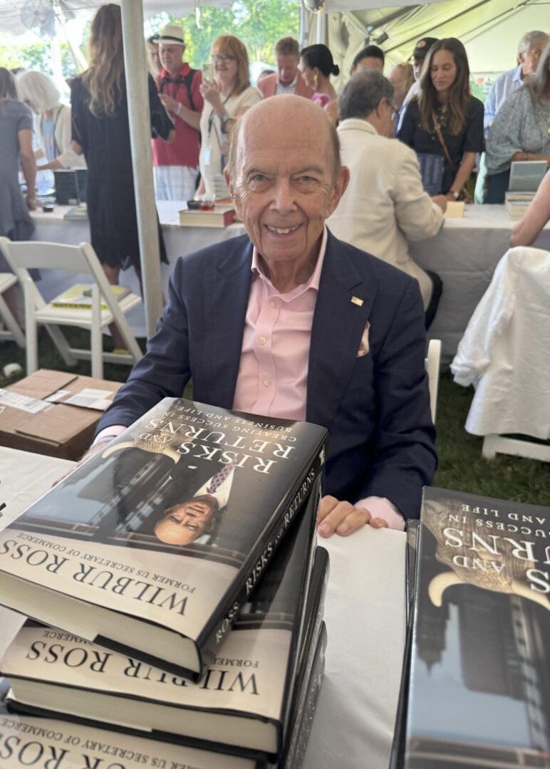 An elderly man at a book signing event with stacks of books.