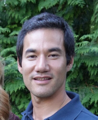 Close-up of a smiling man outdoors with green foliage in the background.