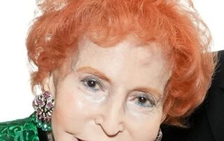 Elderly woman with bright orange hair and vibrant green attire smiling warmly.